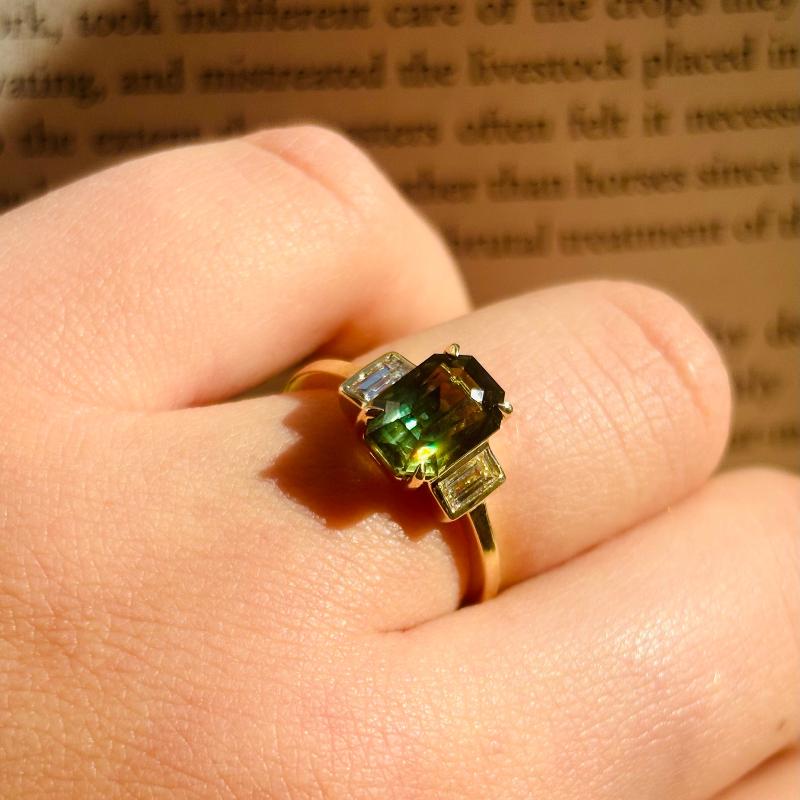 
                  
                    Load image into Gallery viewer, When the sunlight hits these green hues of this emerald cut sapphire and diamond ring, you&amp;#39;ll be obsessed. It has a nice high dome, is cast in 14 kt yellow gold, the center bi-color, emerald cut sapphire is set between 2 baguette cut diamonds.
                  
                