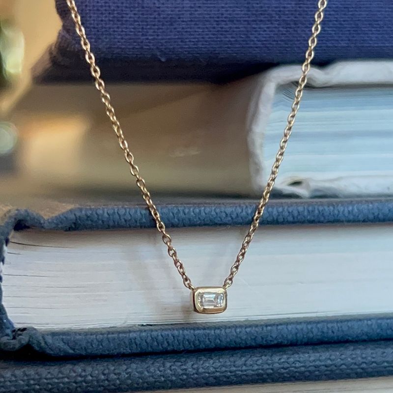 A single baguette diamond bezel set in 14kt yellow gold with blue and white books in the background.