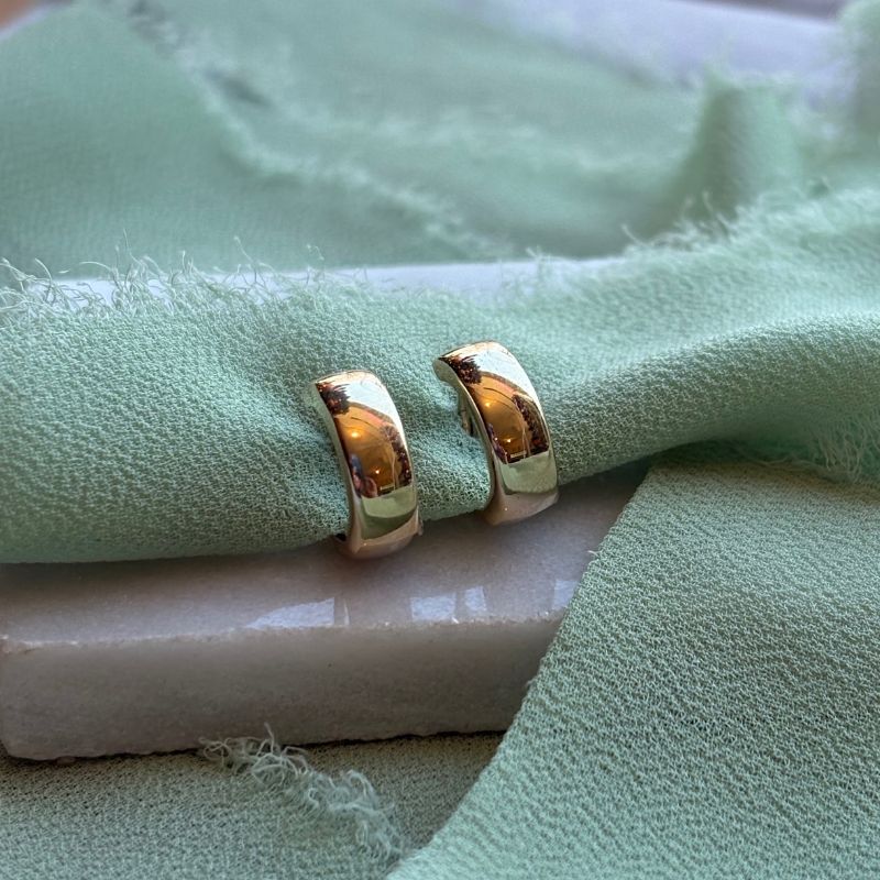 The Piper 14kt yellow gold huggies are shown from the front on a marble tray with green linen draped around.