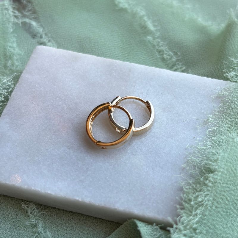 A pair of 14kt yellow gold huggies styled on a marble tray with light green linen draped around