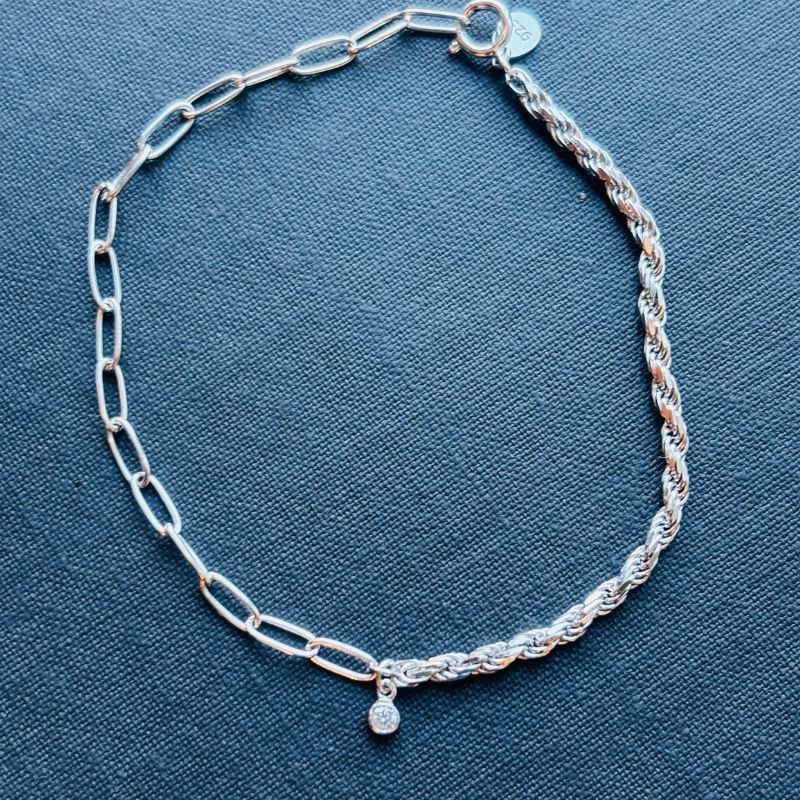 Half paperclip and half rope bracelet joined in the middle by a bezel set round crystal shown on a blue book