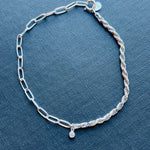 Half paperclip and half rope bracelet joined in the middle by a bezel set round crystal shown on a blue book